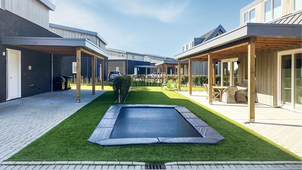 Een moderne woonwijk met een centrale trampoline op een verzorgd gazon, omringd door huizen met overdekte patio's en grote ramen.