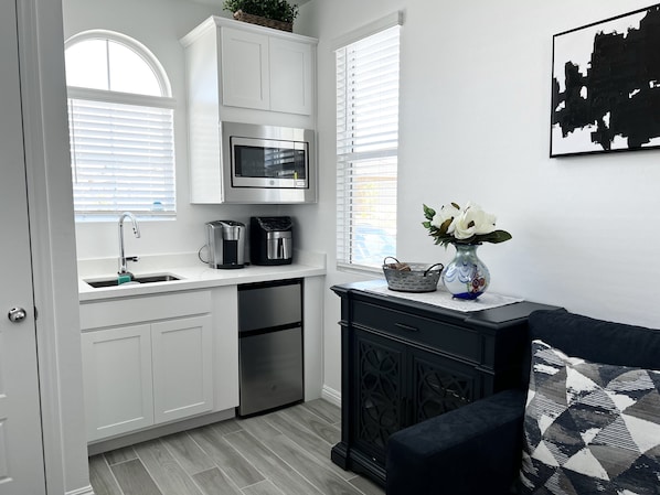 A compact kitchen with white cabinetry, a stainless steel microwave, a coffee maker, and a dishwasher. There is a window with white blinds, a vase with flowers on a black console table, and a black and white patterned pillow on a black sofa.