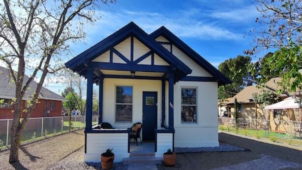 Ein kleines, eingeschossiges Haus mit dunkelblauem Dach und weißen Wänden, mit einer Veranda, zwei Blumentöpfen und einer Bank.