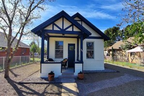 Een klein, gelijkvloers huis met een donkerblauwe dak en witte muren, met een voortuin en een bankje, voorzien van twee plantenpotten.