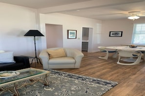 Ein Wohnzimmer mit einer Couch, einem beigefarbenen Sessel, einem Glastisch, einem gemusterten Teppich, einem Esstisch mit Stühlen und einem Deckenventilator.