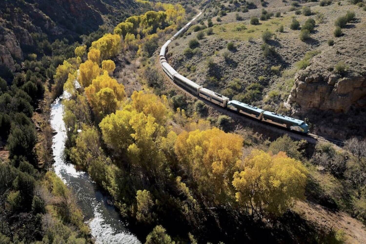 A train traveling through a canyon with a river and yellow foliage.