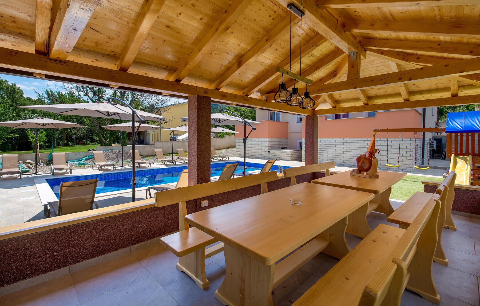 Un espace extérieur aménagé pour les repas, avec des bancs et des tables en bois, une piscine et des parasols.