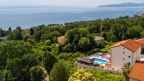 Un barrio residencial con una casa grande que tiene piscina, rodeado de abundante vegetación y vistas al mar.