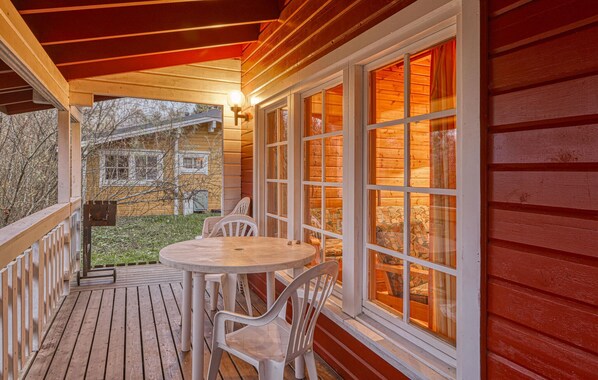 Une terrasse en bois avec une table ronde et des chaises, de grandes fenêtres avec des rideaux, et une vue sur une maison en arrière-plan.