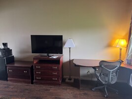 Una habitación de hotel con tocador de madera, televisor de pantalla plana, escritorio con lámpara y silla de oficina de malla.