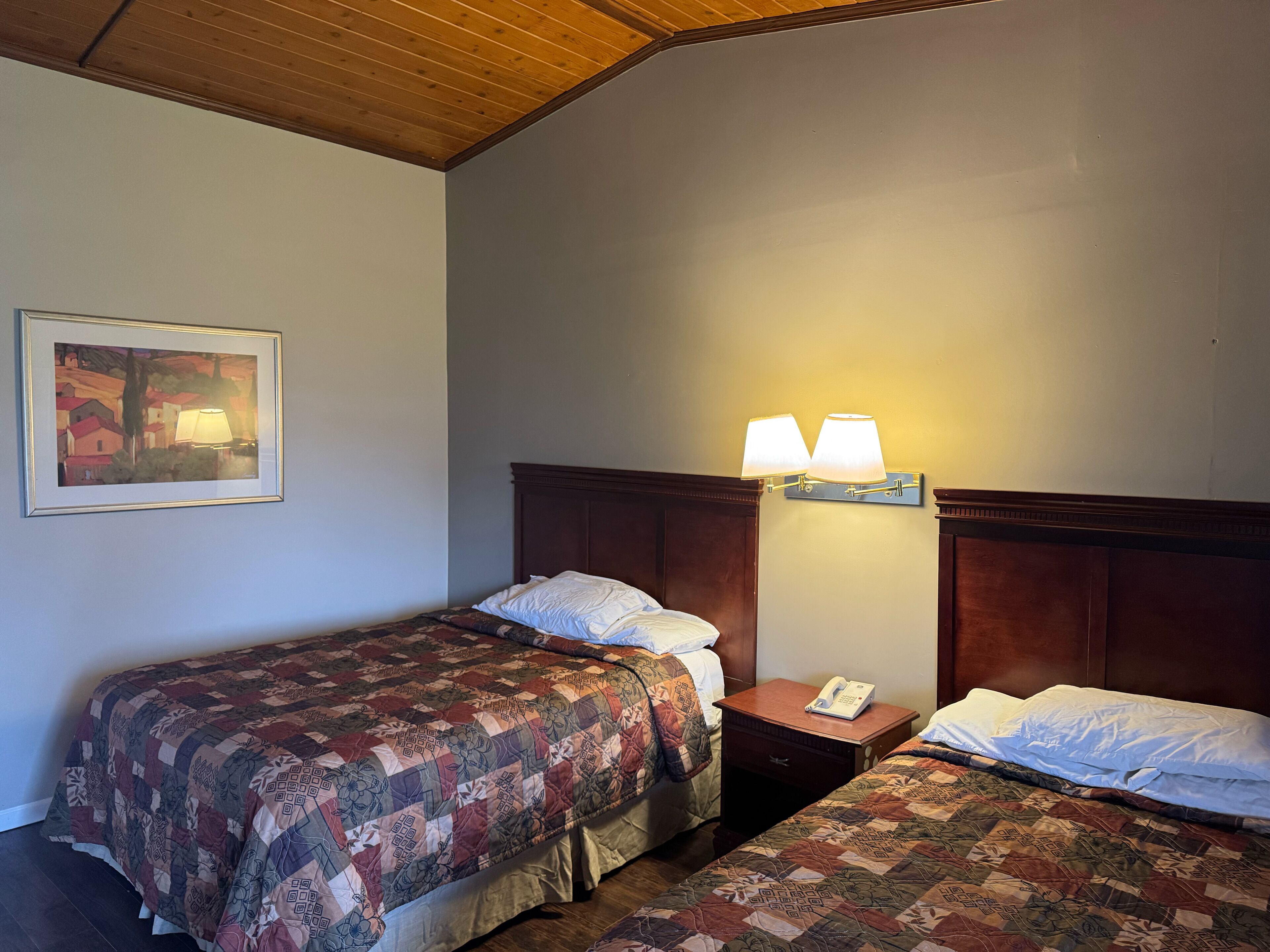Habitación de hotel con dos camas, techo de madera, un cuadro enmarcado en la pared y una mesita de noche con una lámpara.