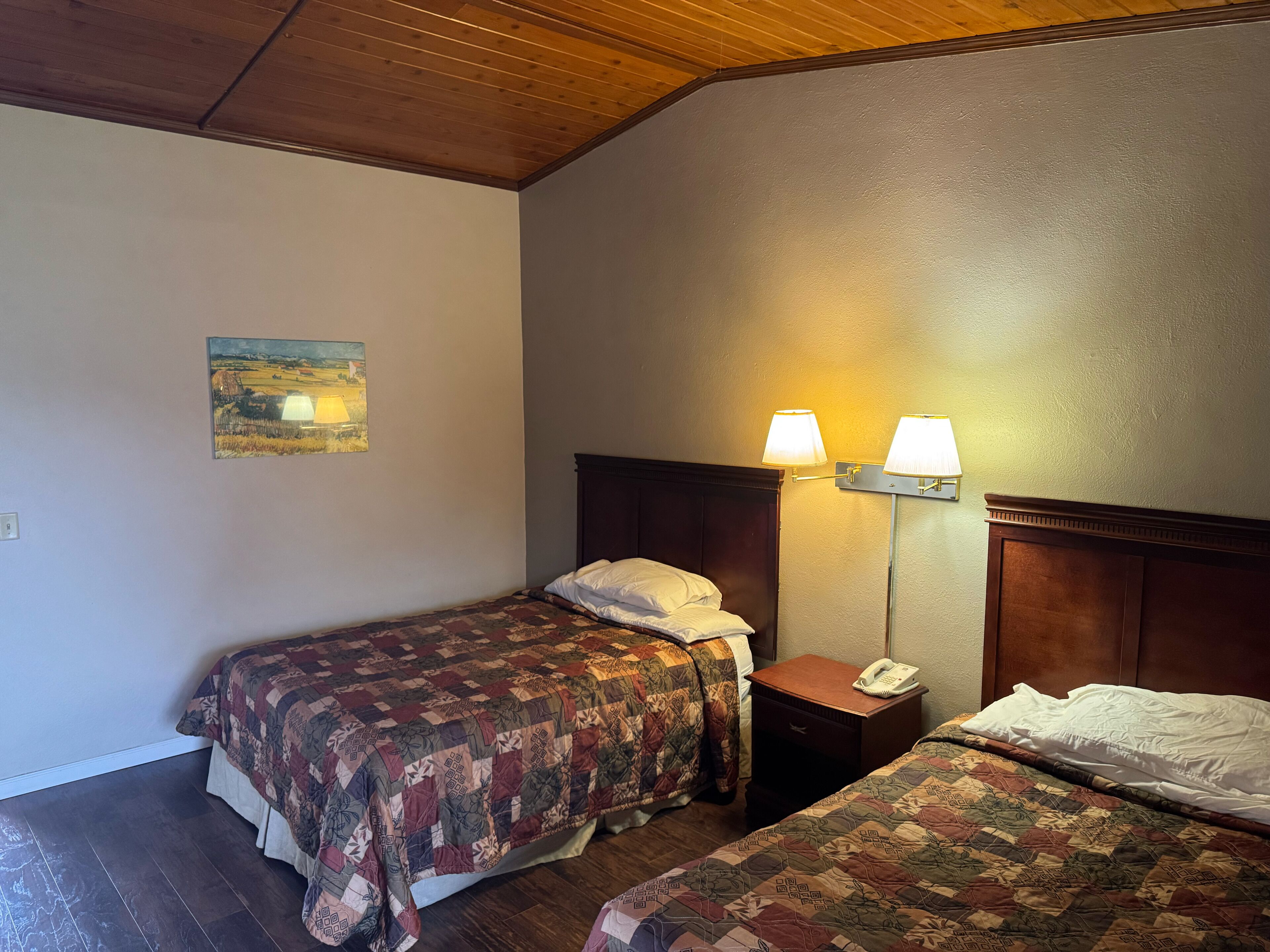 A hotel room with two beds, wooden headboards, a nightstand with a lamp, and a painting on the wall.