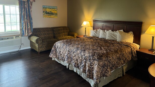 A bedroom with a large bed, two bedside lamps, a headboard, a window with curtains, a painting on the wall, and a patterned sofa.
