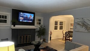 Ein Wohnzimmer mit Kamin, einem darüber angebrachten Fernseher, einer Couch, einem Couchtisch und einem durch einen Bogen sichtbaren Essbereich.