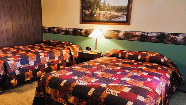Una habitación de hotel con dos camas, cada una cubierta con un edredón colorido, una mesita de noche con una lámpara, y un cuadro de una escena forestal en la pared.