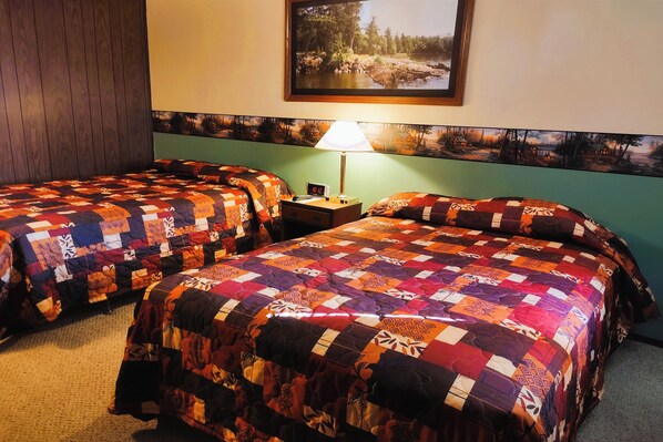 A hotel room with two beds, each covered with a colorful quilt, a nightstand with a lamp, and a painting of a forest scene on the wall. - Love Hotels Voyageur by OYO at International Falls MN (International Falls)