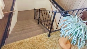 Una escalera con paneles de madera, barandilla negra, peldaños alfombrados y una planta en maceta al pie.