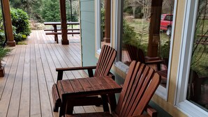 Una terraza de madera con dos sillas y una mesa, con vista a un jardín que tiene un coche rojo al fondo.