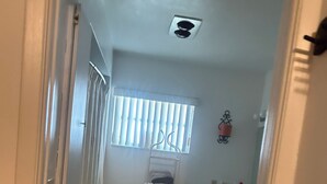 A narrow hallway with a tiled floor, a window with vertical blinds, a metal shelf with folded clothes, a sink, and a mirror.