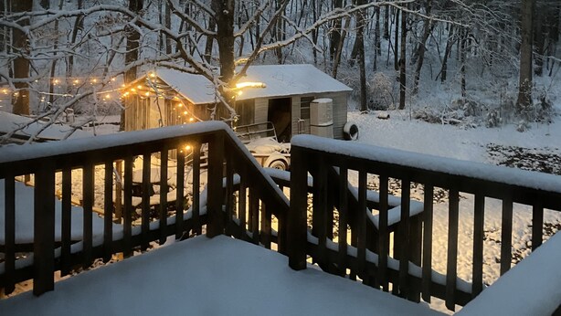 Een houten terras met sneeuw bedekte relingen, een klein huisje met licht en een bebost landschap op de achtergrond.
