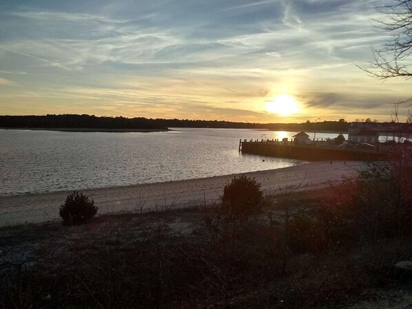 Ein Sonnenuntergang über ruhigem Wasser mit einem Steg, der sich ins Wasser erstreckt, umgeben von spärlicher Vegetation und einer fernen Küstenlinie.