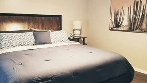 A neatly made bed with a dark blue bedspread and pillows, a wooden headboard, a bedside table with a lamp, and a cactus artwork on the wall. - Hiker's Haven - The Start of a Beautiful, Adventurous and Peaceful Vacation (Sedona)