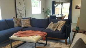 A living room with a blue sofa, an ottoman, and a grey armchair. There is a dining area visible through a glass door. - Hiker's Haven - The Start of a Beautiful, Adventurous and Peaceful Vacation (Sedona)