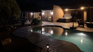 Een zwembadgebied bij nacht met een jacuzzi, loungebedden en een terras met tafels en stoelen.
