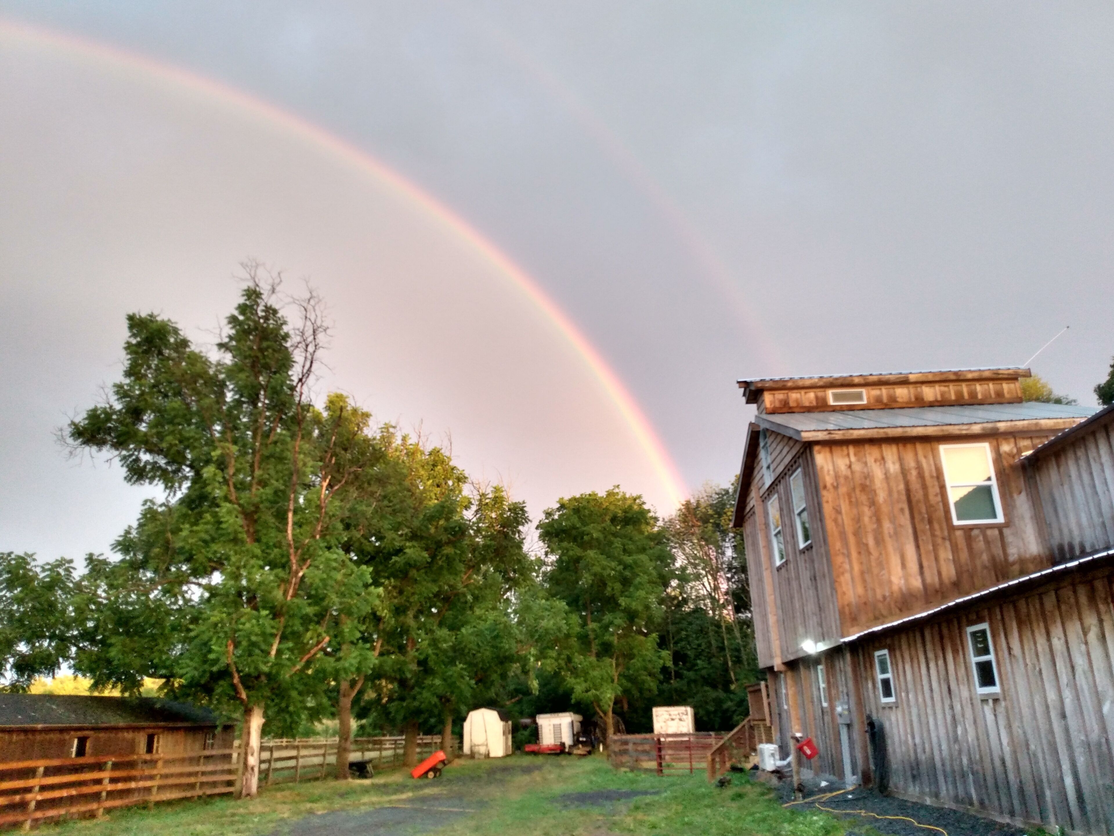 Un bâtiment rustique en bois, doté d’un toit en pente, d’une cour clôturée et d’un arc-en-ciel dans le ciel.