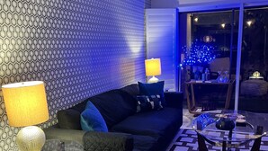 Un salon moderne avec un mur orné d’un motif, un canapé foncé, une table basse en verre et un pouf rond agrémenté de broderies florales.