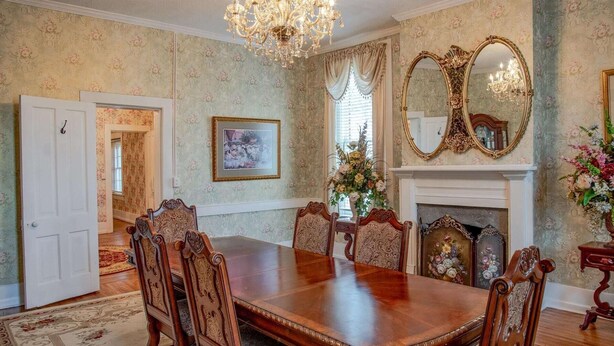 Une salle à manger élégante, avec une grande table en bois, des chaises richement décorées, un lustre, une cheminée et des décorations florales.
