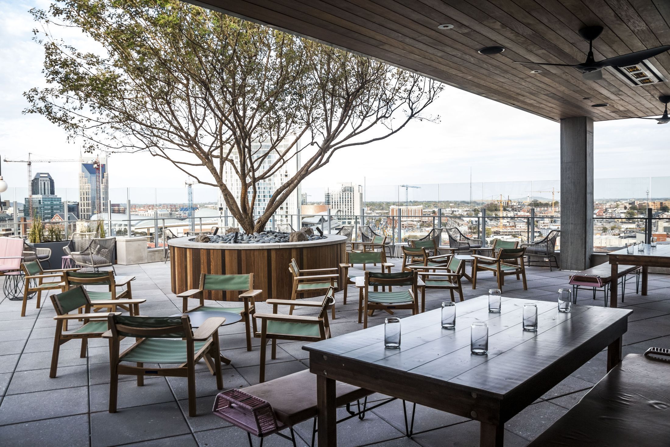 An outdoor seating area with wooden tables and chairs, a large tree, and a view of the cityscape.