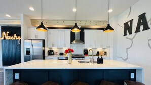 Cocina moderna con armarios blancos, refrigerador de acero inoxidable, una isla central con taburetes, luces colgantes y una pared decorativa con letras.