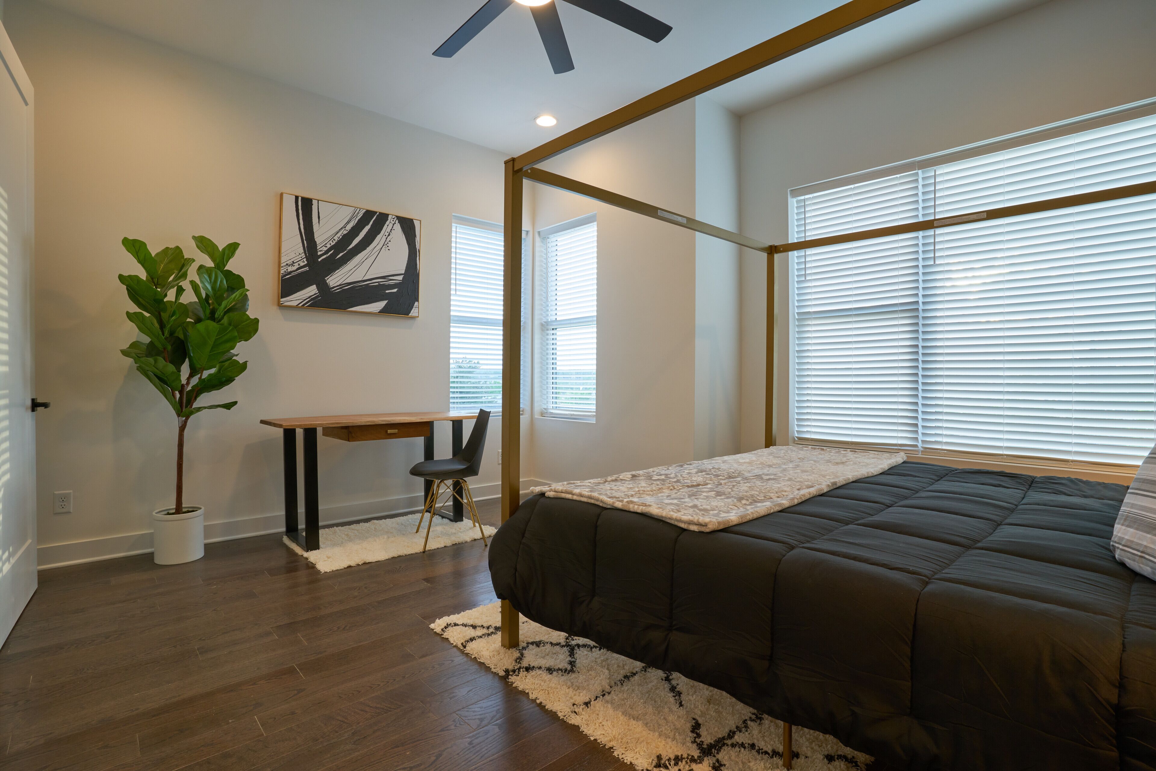 A bedroom with a bed, a desk, a chair, a plant, and a painting on the wall.