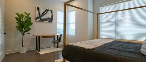 A bedroom with a bed, a desk, a chair, a plant, and a painting on the wall.