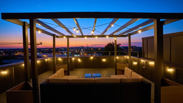 Terraza en la azotea con piso de madera, asientos acolchados e iluminación con luces colgantes.