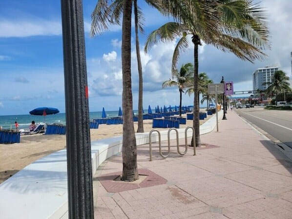 Stroll along the beachfront promenade lined with palm trees, leading to a sandy beach area with umbrellas for relaxation.