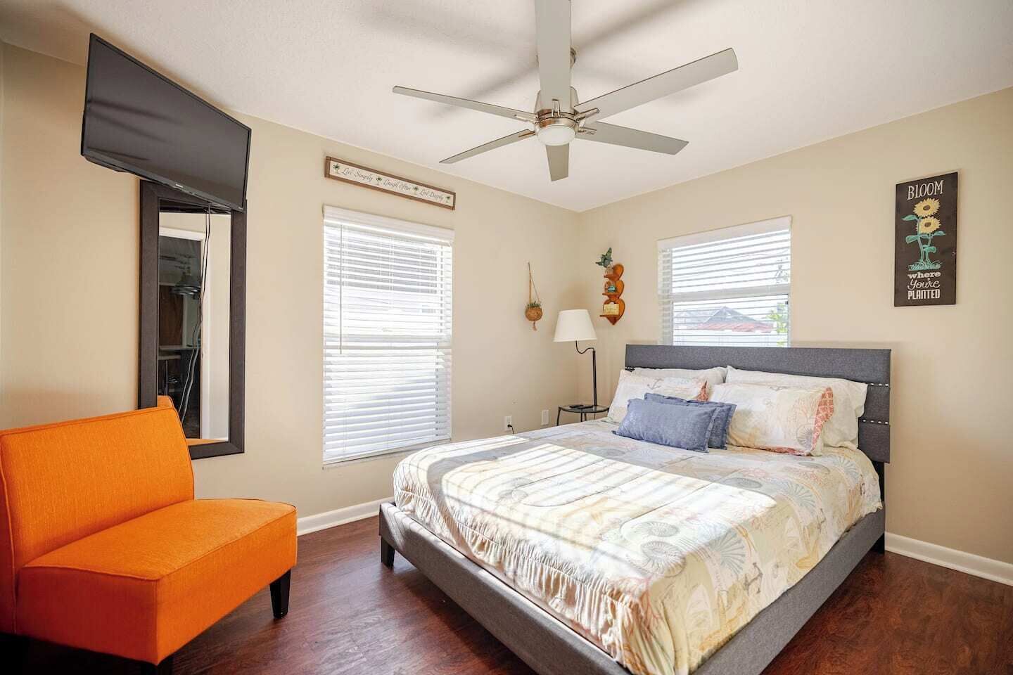 Une chambre à coucher avec un lit, un fauteuil orange, un ventilateur de plafond et un téléviseur fixé au mur.