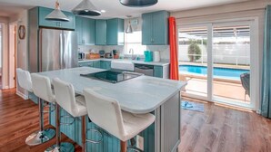 Een moderne keuken met blauwgroene kastjes, een centraal eiland met barkrukken, en uitzicht op een zwembad door grote schuifdeuren.