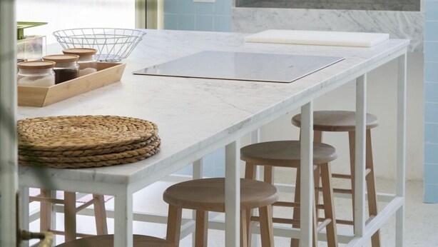 A kitchen with a white marble countertop, wooden stools, and a wire basket with jars.