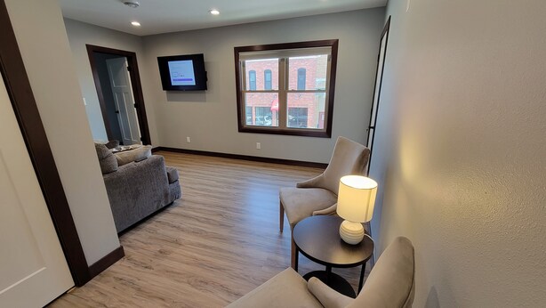 A spacious living room with hardwood flooring, a grey sofa, a beige armchair, and a small table with a lamp. A flat-screen TV is mounted on the wall, and a window provides a view of the outside. - Completely remodeled downtown apartment!  2 large bedrooms! (Cascade)