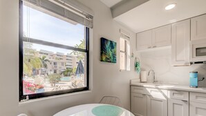 A modern kitchen with white cabinets, a microwave, and a sink. There's a dining table with two chairs and a window with a view of buildings and palm trees. - Poolside Updated Studio Across from the Beach!! (St. Pete Beach)