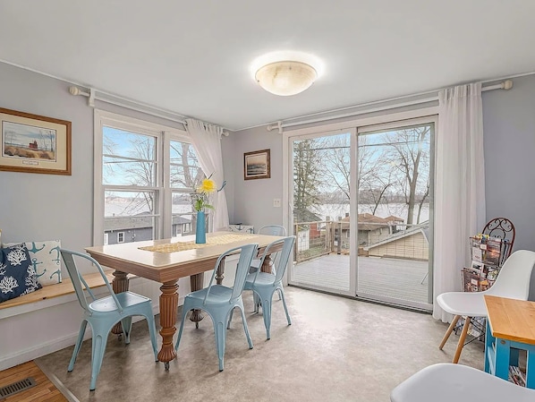 A dining area with a table and chairs, a bench with cushions, and a view through large windows. - Country cottage, Baseline Lake . Come fish, kayak or enjoy a camp fire. (Gobles)