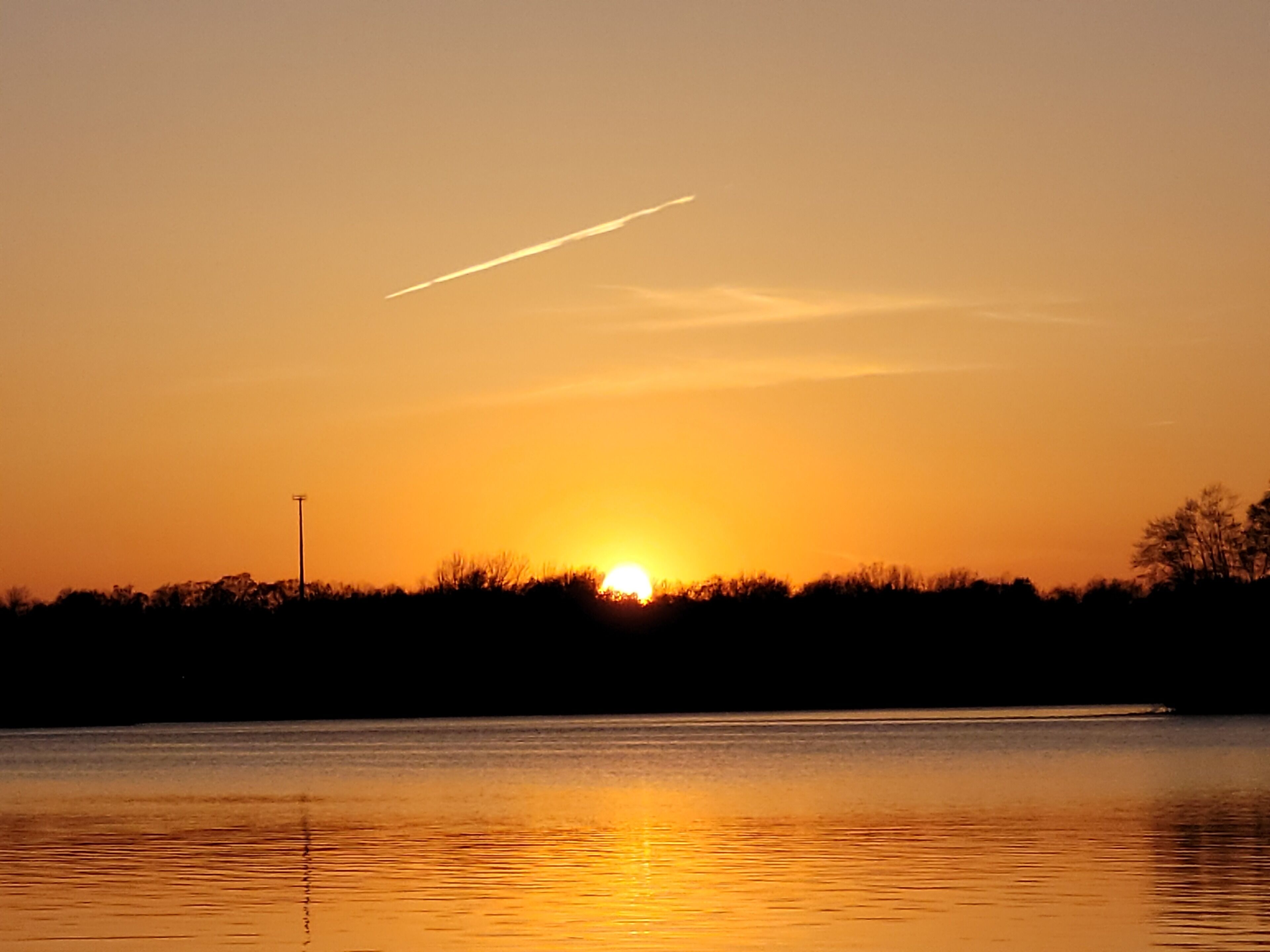 A sunset over a calm body of water with a silhouette of trees and a distant light pole.