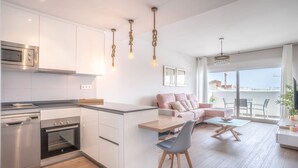 A modern kitchen with white cabinets, stainless steel appliances, and a central island. There is a dining area with a table and chairs, and a living room with a sofa and a balcony with a view. - Stunning Apartment in Estepona, Spain Private Pool (Estepona)