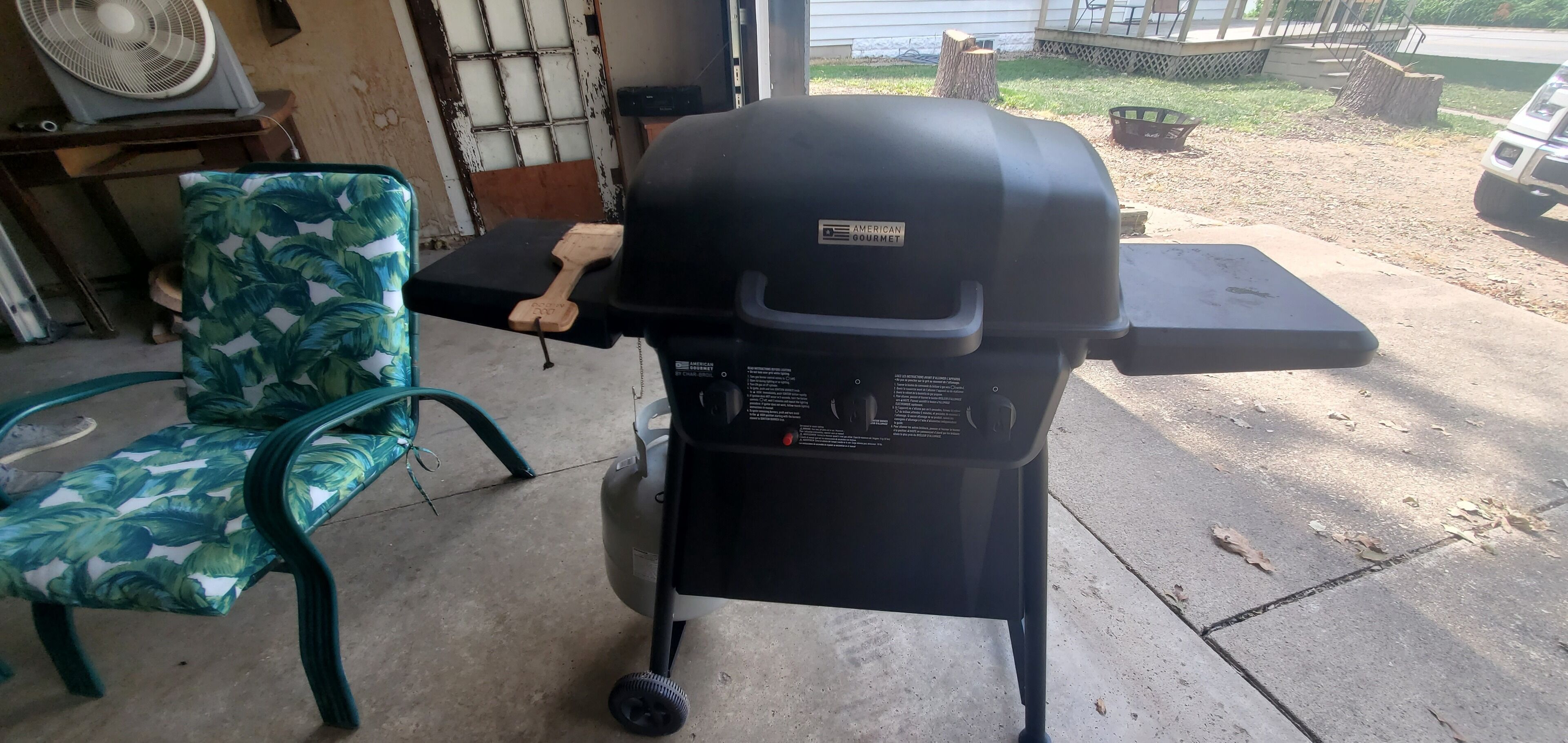 Ein tragbarer Grill mit Aufkleber, ein Stuhl mit Blattmuster und ein Ventilator im Hintergrund.