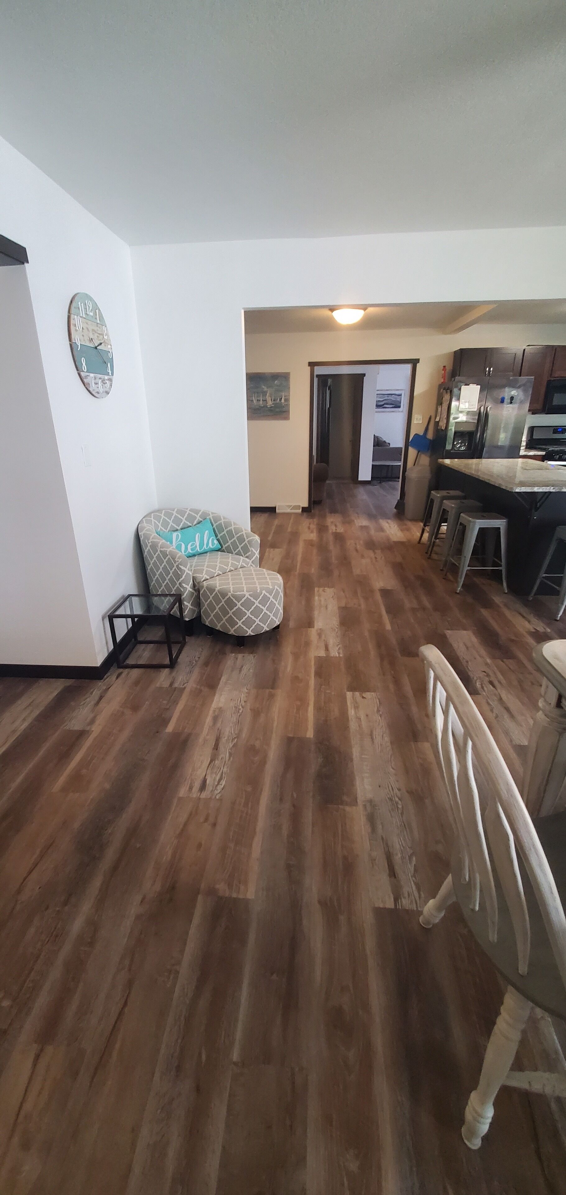 Un'ampia zona giorno con pavimento in legno, una poltrona con motivo decorativo, una zona pranzo e una cucina sullo sfondo, più una porta che conduce in un'altra stanza.