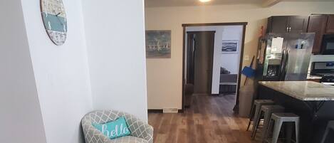 Un'ampia zona giorno con pavimento in legno, una poltrona con motivo decorativo, una zona pranzo e una cucina sullo sfondo, più una porta che conduce in un'altra stanza.