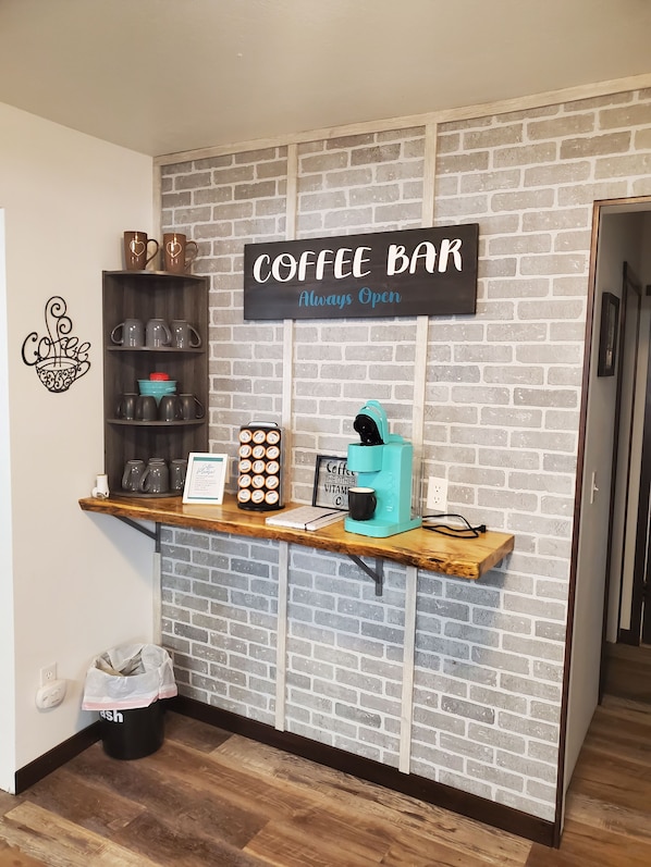 A coffee bar with a sign that reads "Coffee Bar Always Open." There are mugs, a coffee maker, and a trash can visible.