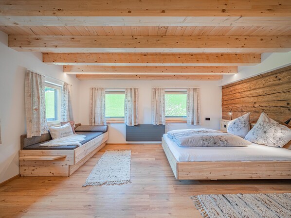 A bedroom with a wooden ceiling, two windows with curtains, a bed with patterned pillows, and a small seating area with a bench.