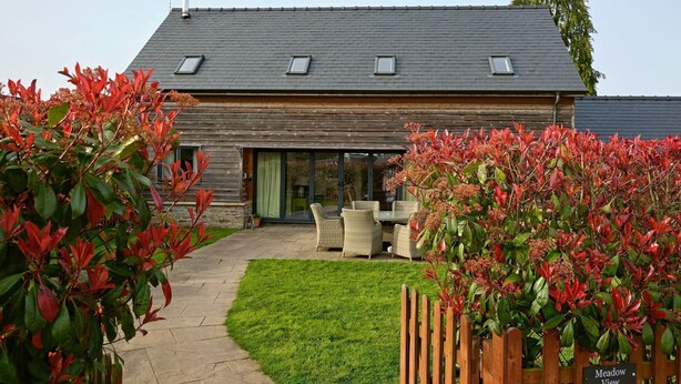 A building with a sloped roof, multiple windows, and a paved entrance area with outdoor furniture. - Estate Lodge -  Luxury Cottages (Leominster)