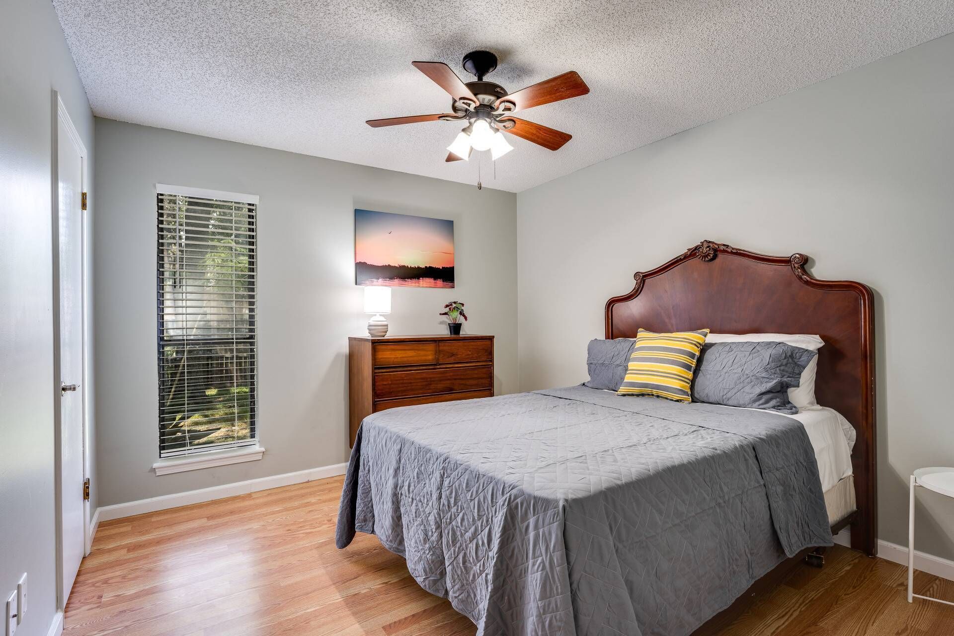 A bedroom with a bed, a dresser, a ceiling fan, a window with blinds, and a painting on the wall.