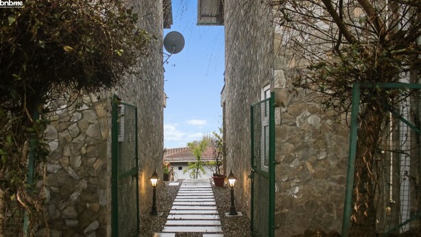 Un couloir aux murs de pierre, avec un chemin central menant à l’entrée d’un bâtiment, de part et d’autre duquel se trouvent des portails verts et des lampes fixées aux murs.