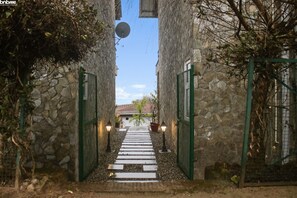 A stone-walled corridor with a central pathway leading to a building entrance, flanked by green gates and wall-mounted lights. - 2B-Mukteshwar-108 by bnbmehomes (Dhari)